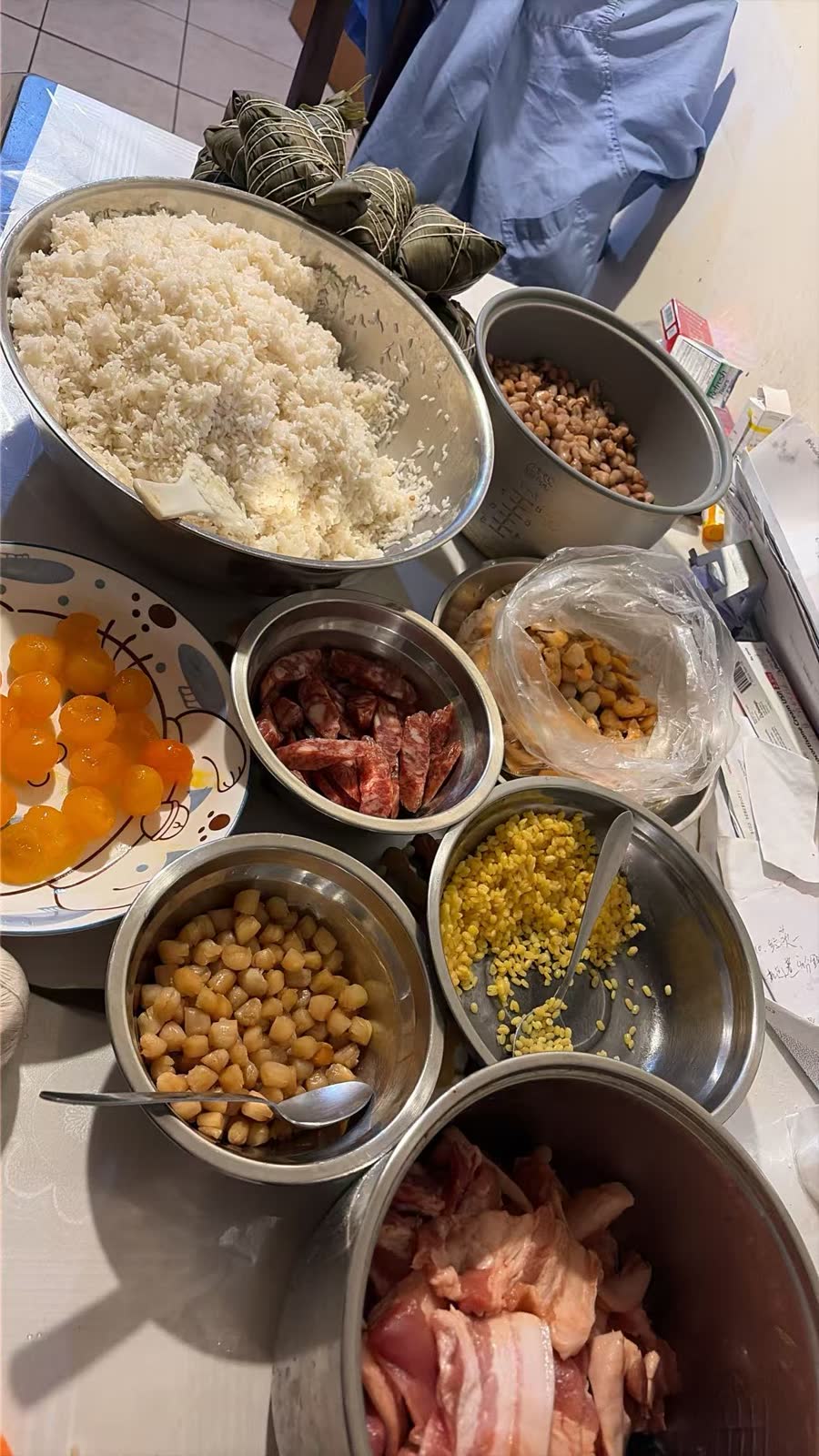 Zongzi mise en place in You Yi’s kitchen — a large bowl of glutinous rice with wrapped bundles behind it, and bowls of salted egg yolks, Chinese sausage, dried shrimp, dried scallops, mung beans, peanuts, and pork belly.